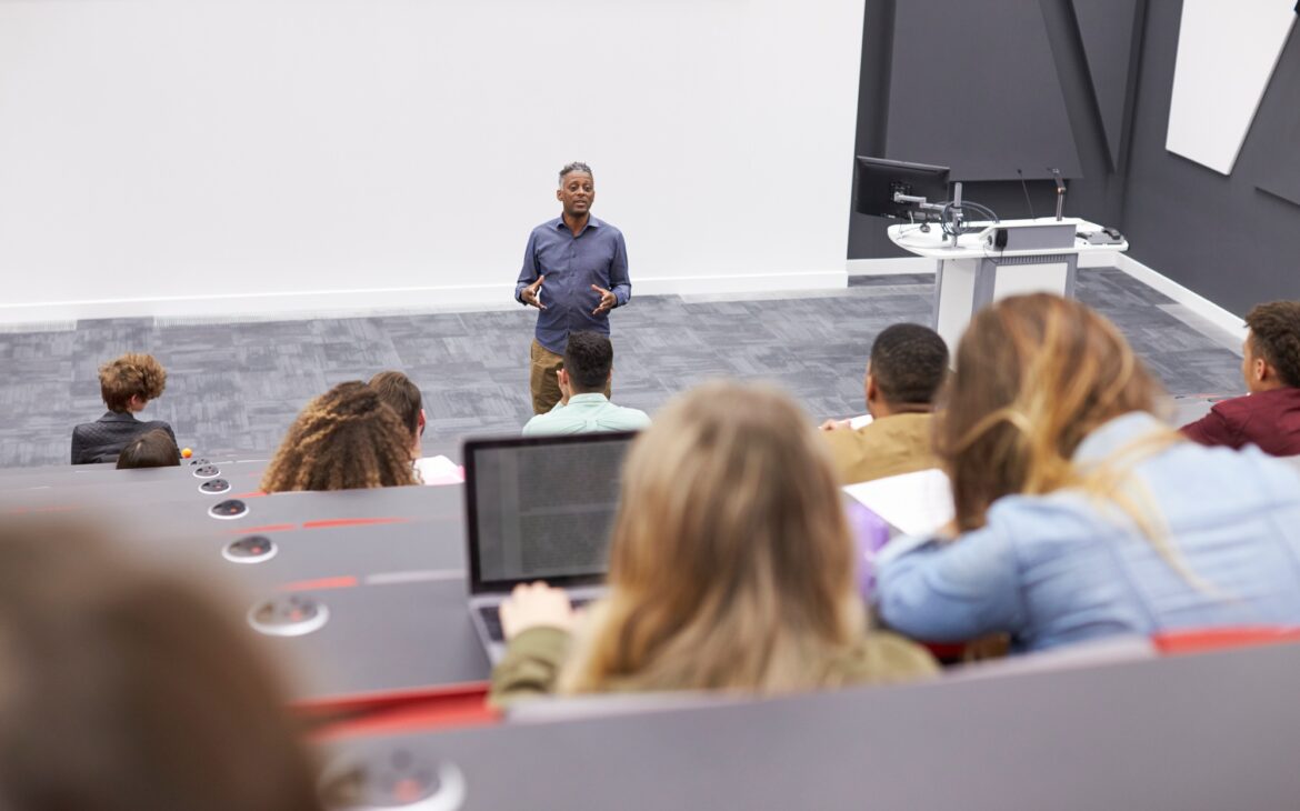 Man lectures students in lecture theatre, back row seat POV