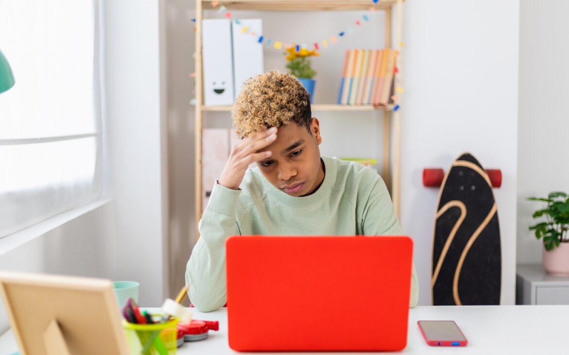 Stressed teenager boy sitting at desk studying on laptop in room