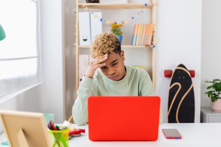 Stressed teenager boy sitting at desk studying on laptop in room
