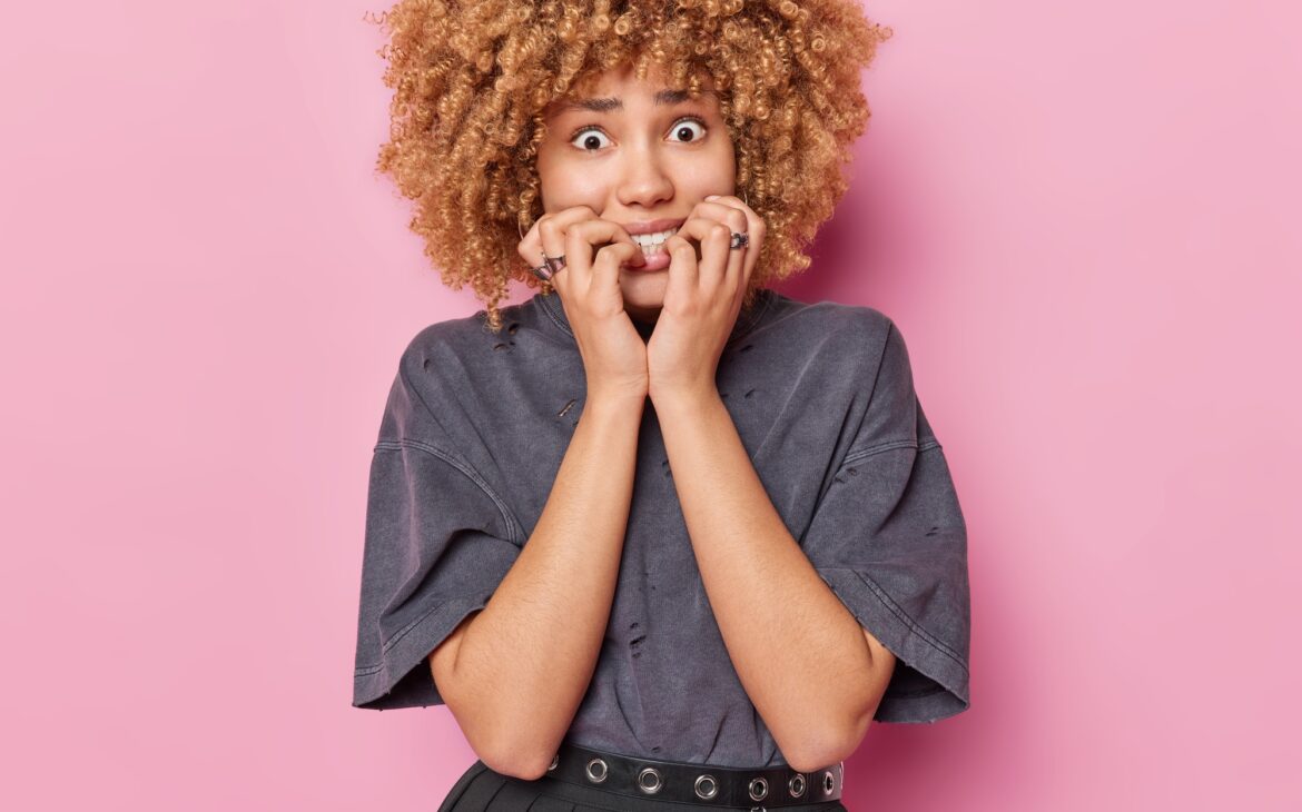 Waist up shot of nervous curly haired woman bite finger nails feels nervous looks anxious at camera