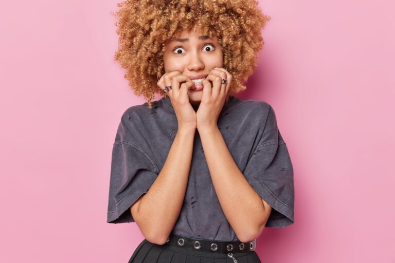 Waist up shot of nervous curly haired woman bite finger nails feels nervous looks anxious at camera